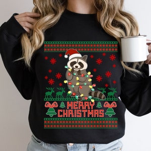 May include: Black Christmas sweater with a raccoon wearing a Santa hat and holding Christmas lights. The sweater has a green and red pattern and says "Merry Christmas".
