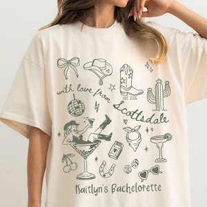 May include: Cream-colored oversized t-shirt with a green graphic design. The design includes a cowboy hat, boots, cactus, and a woman in a martini glass. Text reads "with love from Scottsdale" and "Kaitlyn's Bachelorette".