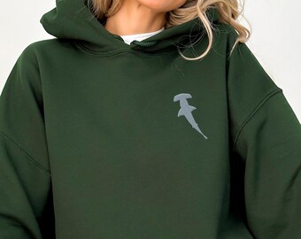 May include: A dark green hoodie with a kangaroo pocket and a drawstring hood. The hoodie features a small, grey hammerhead shark graphic on the upper left chest. The person in the image is wearing blue jeans.