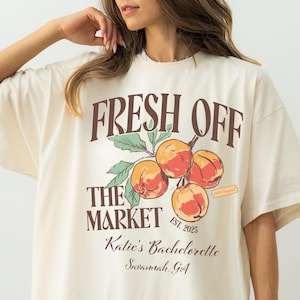 May include: Cream-colored t-shirt with the text "FRESH OFF THE MARKET" above a peach illustration. Below the illustration, the text reads "Katie's Bachelorette Savannah, GA". The shirt has short sleeves and a relaxed fit.
