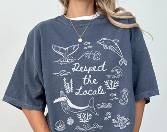 May include: A dark blue t-shirt with a white graphic design featuring a whale, dolphins, and sea life. The text on the shirt reads "Respect the Locals".