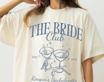 May include: Cream-colored t-shirt with the text "THE BRIDE Club" in blue, along with a graphic of two martini glasses. Below the graphic is "Reagan's Bachelorette, Napa Valley, CA" and the year "2025".
