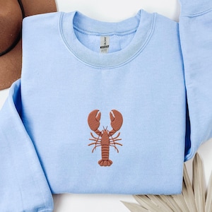 Pode incluir: Uma camisa de moletom azul claro com um lagosta bordada marrom na frente.