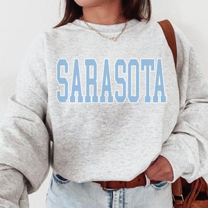 May include: A light gray sweatshirt with the word "SARASOTA" in light blue block letters across the chest. The sweatshirt has long sleeves and a crew neck. The person is wearing light blue jeans and a brown belt.