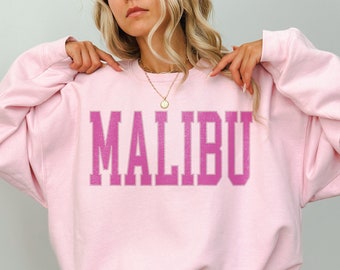Op de afbeelding: Een roze sweatshirt met het woord "MALIBU" in roze op de voorkant. Het sweatshirt is oversized en heeft een ronde hals.