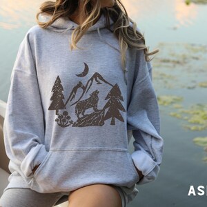 Puede incluir: Una sudadera con capucha gris con un estampado gr&aacute;fico marr&oacute;n de un lobo aullando a la luna en un entorno de bosque. El gr&aacute;fico incluye &aacute;rboles, monta&ntilde;as y una luna creciente.