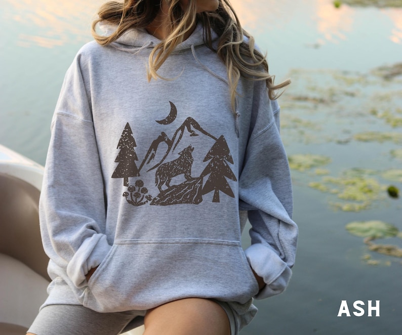 Puede incluir: Una sudadera con capucha gris con un estampado gr&aacute;fico marr&oacute;n de un lobo aullando a la luna en un entorno de bosque. El gr&aacute;fico incluye &aacute;rboles, monta&ntilde;as y una luna creciente.
