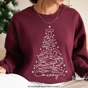 K&ouml;nnte beinhalten: Burgunderfarbenes Sweatshirt mit einer wei&szlig;en Grafik eines Weihnachtsbaums, der aus verschiedenen Haiarten besteht. Das Wort "merry" ist unten in Schreibschrift geschrieben. Das Design beinhaltet Sterne und einen Stern an der Spitze des Baumes.