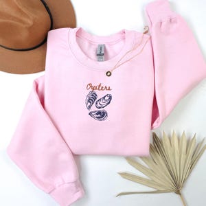 Peut inclure: Sweat-shirt rose &agrave; col rond avec le mot "Oysters" en texte brun et un graphique de trois hu&icirc;tres en bleu.