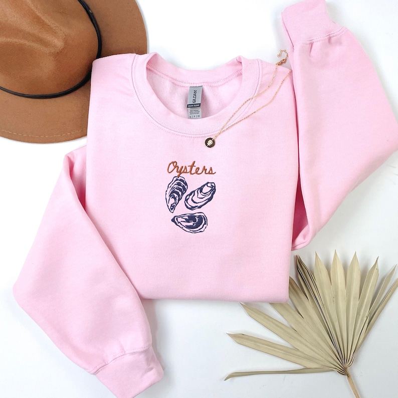 Peut inclure: Sweat-shirt rose &agrave; col rond avec le mot "Oysters" en texte brun et un graphique de trois hu&icirc;tres en bleu.