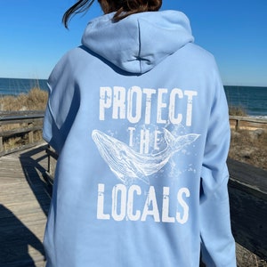 Może przedstawiać: Jasnoniebieska bluza z kapturem z napisem "PROTECT THE LOCALS" i białą grafiką wieloryba na plecach. Bluza z kapturem jest wykonana z miękkiego materiału i ma kaptur. Tło przedstawia plażę i morze.