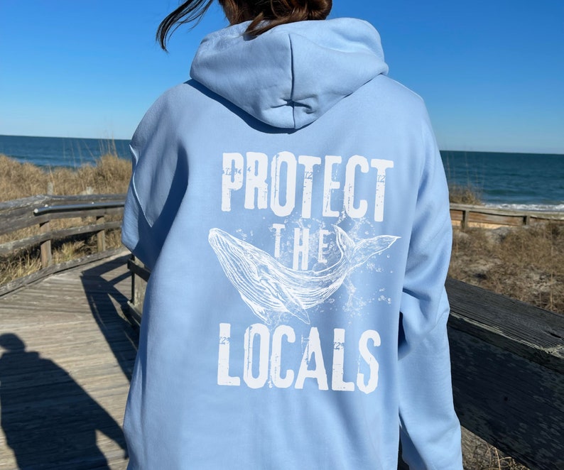 Może przedstawiać: Jasnoniebieska bluza z kapturem z napisem "PROTECT THE LOCALS" i białą grafiką wieloryba na plecach. Bluza z kapturem jest wykonana z miękkiego materiału i ma kaptur. Tło przedstawia plażę i morze.