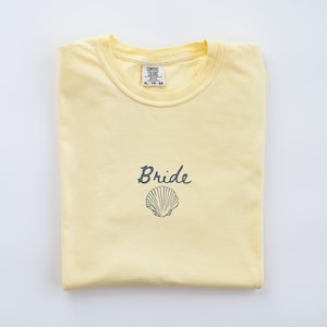 May include: A pale yellow short-sleeve t-shirt featuring the word Bride in dark gray cursive script above a dark gray line-art illustration of a seashell. The inner neck label displays the brand name Comfort Colors and the size XL-TG-EG.
