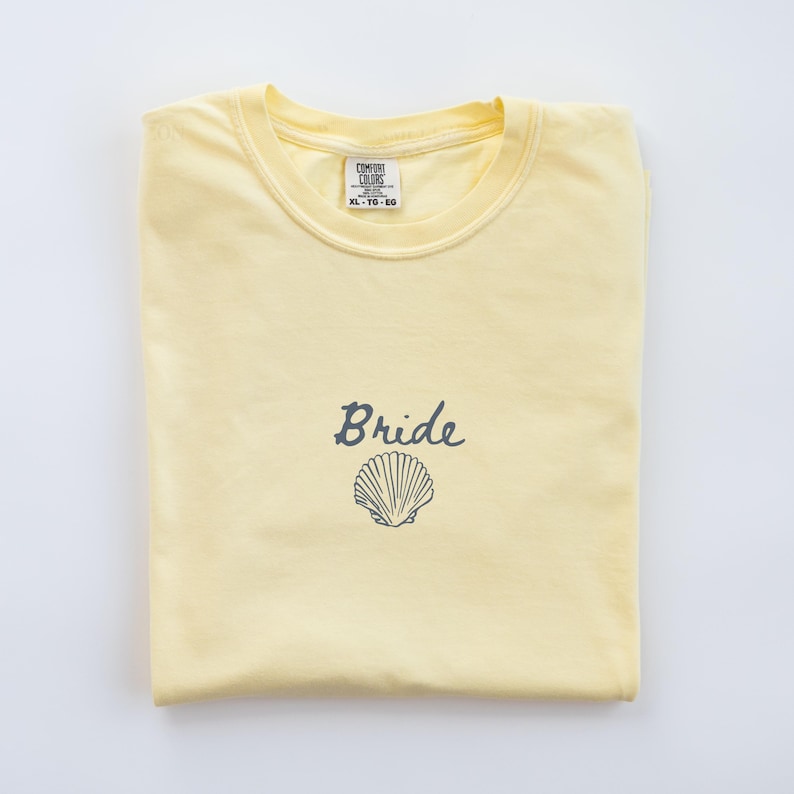 May include: A pale yellow short-sleeve t-shirt featuring the word Bride in dark gray cursive script above a dark gray line-art illustration of a seashell. The inner neck label displays the brand name Comfort Colors and the size XL-TG-EG.