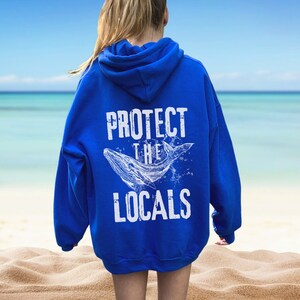 Może przedstawiać: Kr&oacute;lewska niebieska bluza z kapturem z napisem "PROTECT THE LOCALS" i białą ilustracją wieloryba. Bluza ma kaptur i długie rękawy. Tło przedstawia scenę na plaży.
