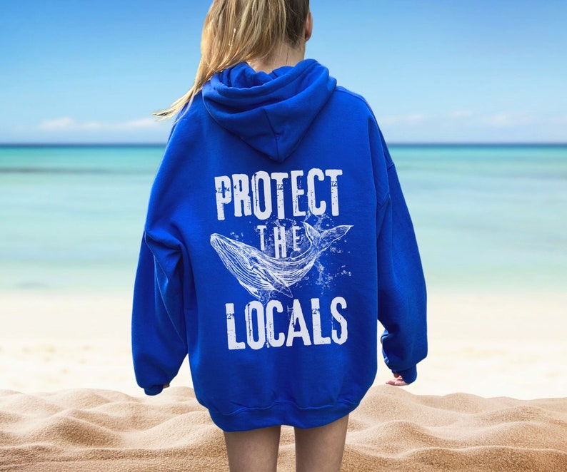 Może przedstawiać: Kr&oacute;lewska niebieska bluza z kapturem z napisem "PROTECT THE LOCALS" i białą ilustracją wieloryba. Bluza ma kaptur i długie rękawy. Tło przedstawia scenę na plaży.