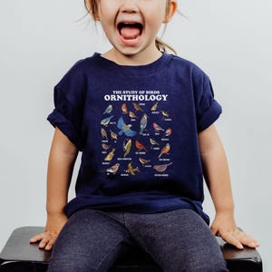May include: A young child wearing a navy blue t-shirt with a white graphic of various birds and the text "THE STUDY OF BIRDS ORNITHOLOGY".