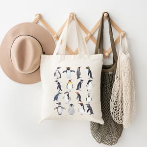 May include: A beige tote bag with a penguin illustration hangs on a wooden wall rack, alongside a tan hat and two other bags. The tote bag features various penguin species in a watercolor style, with black, white, and yellow details. The background is a plain white wall.