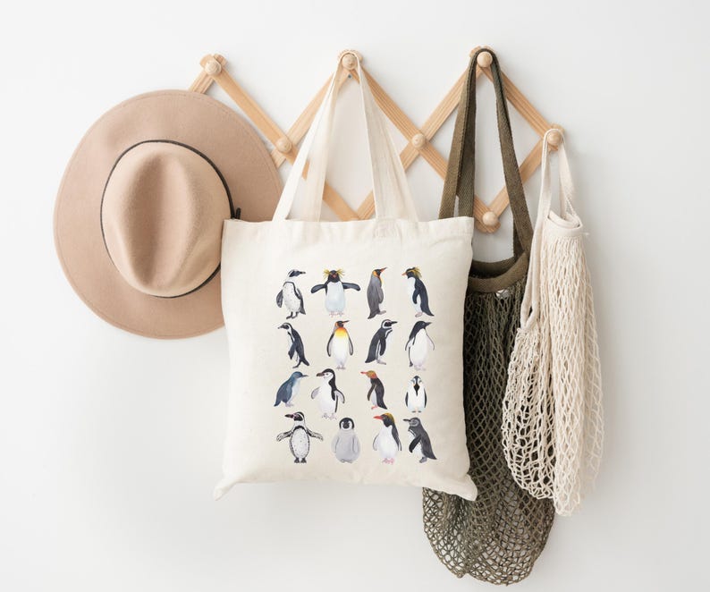 May include: A beige tote bag with a penguin illustration hangs on a wooden wall rack, alongside a tan hat and two other bags. The tote bag features various penguin species in a watercolor style, with black, white, and yellow details. The background is a plain white wall.