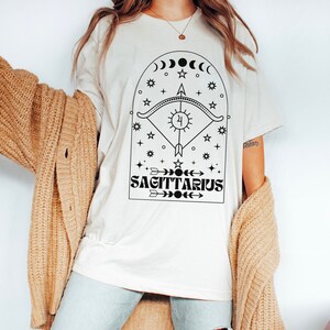 May include: White t-shirt with a black graphic design featuring a Sagittarius zodiac symbol with an arrow, sun, moon, and stars. The text "SAGITTARIUS" is printed below the symbol.