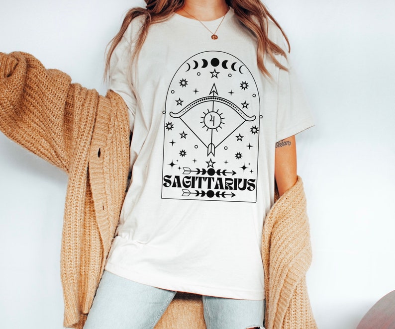May include: White t-shirt with a black graphic design featuring a Sagittarius zodiac symbol with an arrow, sun, moon, and stars. The text "SAGITTARIUS" is printed below the symbol.