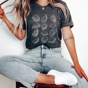 May include: A woman wearing a dark gray t-shirt with a white outline of oysters printed on it. She is wearing light blue jeans and white sneakers. The text "Nine Oceans" is tattooed on her arm.