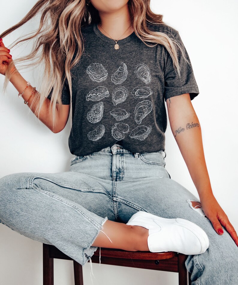 May include: A woman wearing a dark gray t-shirt with a white outline of oysters printed on it. She is wearing light blue jeans and white sneakers. The text "Nine Oceans" is tattooed on her arm.