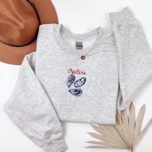 Peut inclure: Un sweat-shirt gris avec le mot "Oysters" brod&eacute; en orange sur le devant. Trois illustrations d'hu&icirc;tres bleues et blanches sont situ&eacute;es sous le mot.