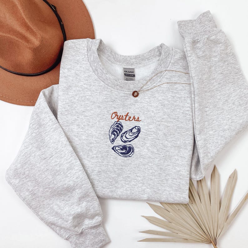Peut inclure: Un sweat-shirt gris avec le mot "Oysters" brod&eacute; en orange sur le devant. Trois illustrations d'hu&icirc;tres bleues et blanches sont situ&eacute;es sous le mot.