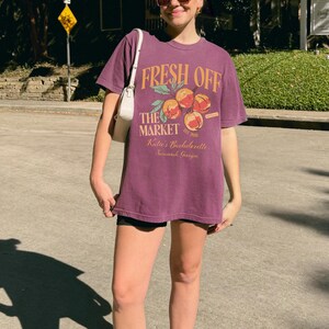 May include: A purple t-shirt with the text "FRESH OFF THE MARKET" in orange, along with a peach graphic. The shirt also includes the text "Katie's Bachelorette Savannah, Georgia". The shirt is worn with black shorts and a white purse.