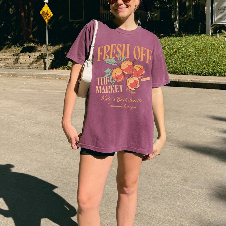 May include: A purple t-shirt with the text "FRESH OFF THE MARKET" in orange, along with a peach graphic. The shirt also includes the text "Katie's Bachelorette Savannah, Georgia". The shirt is worn with black shorts and a white purse.