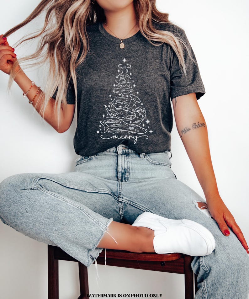 K&ouml;nnte beinhalten: Dunkelgraues T-Shirt mit einem wei&szlig;en Hai-Design in Form eines Weihnachtsbaums. Das Wort "merry" steht darunter. Das T-Shirt besteht aus weichem Material. Ein l&auml;ssiges, festliches Oberteil.