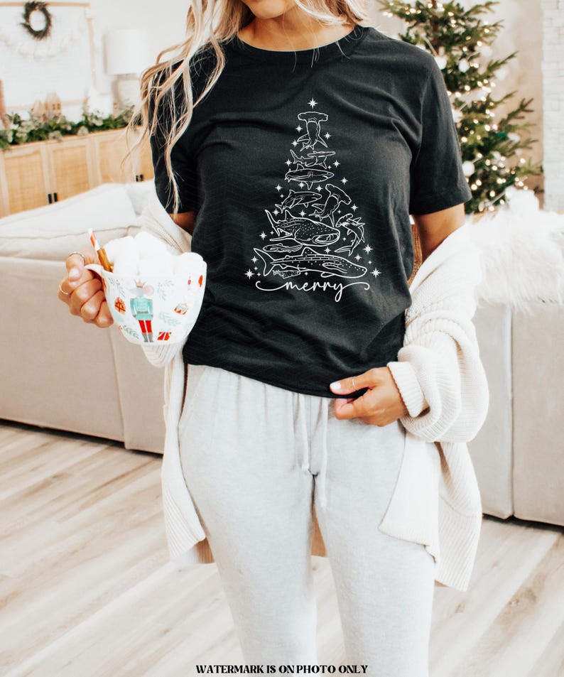 K&ouml;nnte beinhalten: Schwarzes T-Shirt mit einem wei&szlig;en Grafikdesign eines Weihnachtsbaums aus verschiedenen Haiarten. Das Wort "merry" ist darunter in Schreibschrift geschrieben. Das T-Shirt wird mit grauer Jogginghose kombiniert.