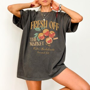 May include: Dark gray t-shirt with the text "FRESH OFF THE MARKET" in gold and a peach graphic. The shirt also includes the text "Katie's Bachelorette Savannah, GA" and "EST. 2023".