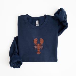 Pode incluir: Uma camisola azul marinho com um lagosta bordada a castanho na frente.