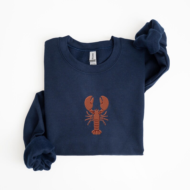 Pode incluir: Uma camisola azul marinho com um lagosta bordada a castanho na frente.