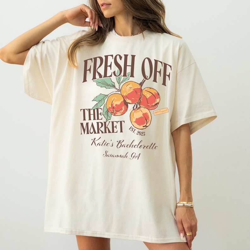 May include: Cream-colored t-shirt with the text "FRESH OFF THE MARKET" above a peach illustration. Below the illustration, the text reads "Katie's Bachelorette Savannah, GA". The shirt has short sleeves and a relaxed fit.