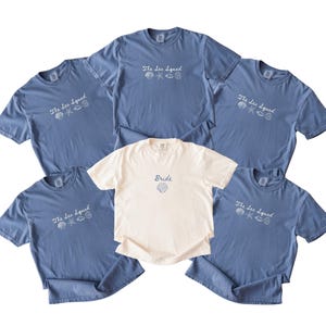 May include: Five blue t-shirts and one white t-shirt. The blue shirts feature the text "The Sea Squad" in white with small shell and starfish graphics. The white shirt features the text "Bride" in blue with a single shell graphic below it.