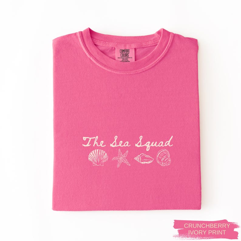 May include: A bright pink short-sleeve t-shirt featuring the white text The Sea Squad above a line of four white illustrations of seashells and a starfish. The inner neck label displays the Comfort Colors brand logo and text.