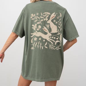 Pu&ograve; includere: Una maglietta verde oversize con una stampa bianca di due conigli e disegni floreali sul retro.
