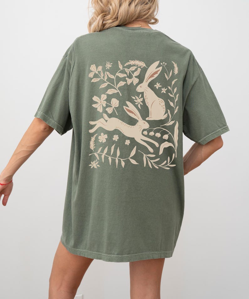 Pu&ograve; includere: Una maglietta verde oversize con una stampa bianca di due conigli e disegni floreali sul retro.