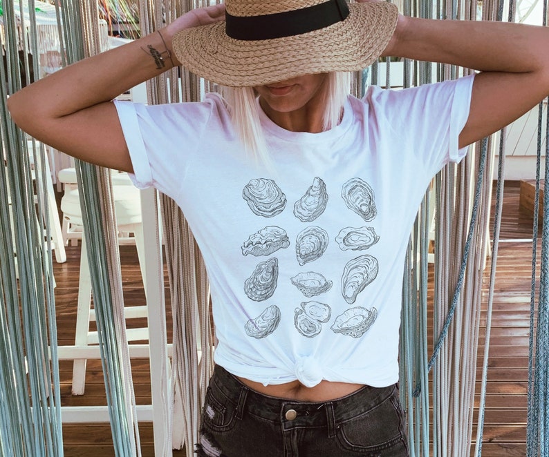 May include: White t-shirt with a graphic print of various oysters. The shirt is worn by a person with a straw hat.
