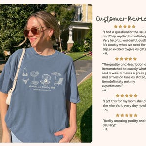 May include: A slate blue short-sleeved t-shirt featuring a white line-art graphic of a bikini, cocktail, pearl in a shell, umbrella, and beach chair. The text reads Seashells and Wedding Bells and OLIVIA'S BACHELORETTE in a cursive and block font.