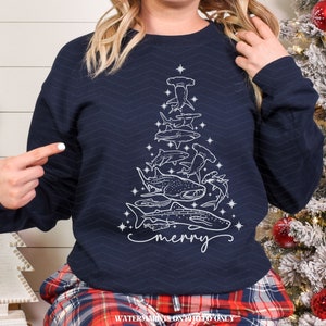 K&ouml;nnte beinhalten: Marineblaues Sweatshirt mit einem wei&szlig;en Weihnachtsbaum-Design aus verschiedenen Haiarten. Das Wort "merry" ist in Schreibschrift geschrieben. Das Sweatshirt hat einen Rundhalsausschnitt und lange &Auml;rmel.