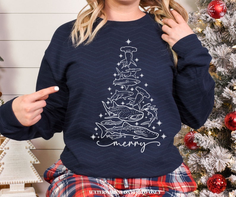 K&ouml;nnte beinhalten: Marineblaues Sweatshirt mit einem wei&szlig;en Weihnachtsbaum-Design aus verschiedenen Haiarten. Das Wort "merry" ist in Schreibschrift geschrieben. Das Sweatshirt hat einen Rundhalsausschnitt und lange &Auml;rmel.