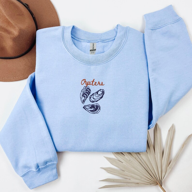 Peut inclure: Un sweat-shirt bleu clair avec le mot "Oysters" brod&eacute; en orange sur le devant. Trois coquilles d'hu&icirc;tres stylis&eacute;es sont brod&eacute;es sous le mot.