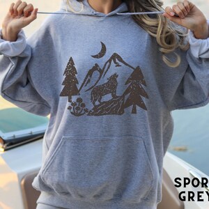 Puede incluir: Una sudadera con capucha gris con un lobo marr&oacute;n aullando a la luna en una escena de bosque. El texto "SPORT GREY" est&aacute; impreso en la esquina inferior derecha de la imagen.