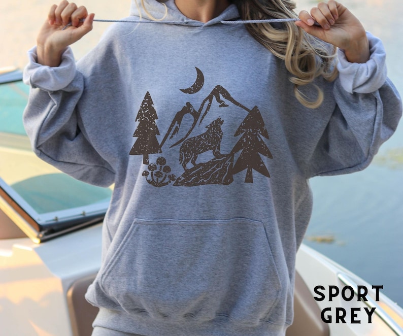 Puede incluir: Una sudadera con capucha gris con un lobo marr&oacute;n aullando a la luna en una escena de bosque. El texto "SPORT GREY" est&aacute; impreso en la esquina inferior derecha de la imagen.