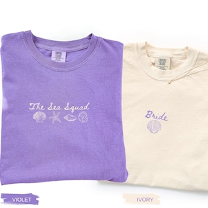 May include: A violet t-shirt with white embroidery reading The Sea Squad above a row of four seashells and a starfish, and an ivory t-shirt with violet embroidery reading Bride above a single seashell. Both shirts feature Comfort Colors labels.