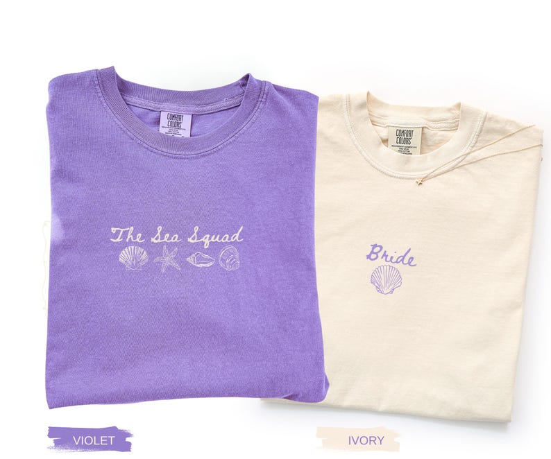May include: A violet t-shirt with white embroidery reading The Sea Squad above a row of four seashells and a starfish, and an ivory t-shirt with violet embroidery reading Bride above a single seashell. Both shirts feature Comfort Colors labels.
